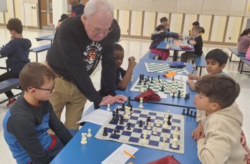 An instructor teaches two young children chess in the foreground, and several pairs of children play chess in the background.